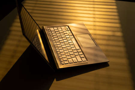 A woman is typing on a laptop keyboard on a white table. The shadow from the blinds falls on the...