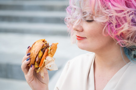Caucasian woman with curly colored hair eating burger. Bad eating habits and love of fast foodの写真素材