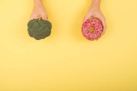 Comparison of eating habits. Woman holding broccoli and donut on a yellow background. Cope space.の写真素材