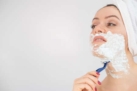 A woman with a white terry towel on her head and with shaving foam on her face shaves on a white backgroundの写真素材
