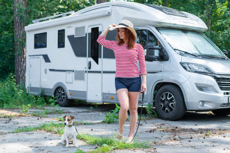 Caucasian red-haired woman in a straw hat and striped sweater walks with a small dog in front of a mobile home. Travel in a camper van with your petの写真素材