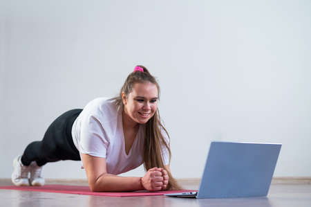A plump young woman stands in a plank and watches an online fitness lesson on a laptop. Distance...