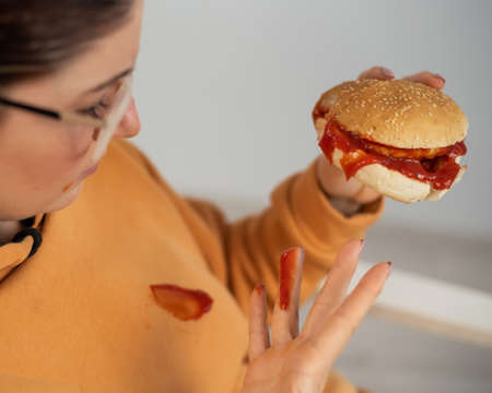 Caucasian young woman eating burger and getting dirty in ketchup. Sloppy girlの写真素材