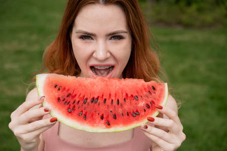 Close-up portrait of red-haired young woman with braces eating watermelon outdoors