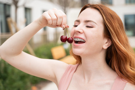 Beautiful young red-haired woman with braces on her teeth eats sweet cherries in the summer outdoorsの写真素材
