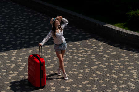 Happy caucasian young woman in hat and shorts holding a large red suitcase outdoors.の写真素材