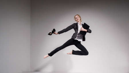 Graceful barefoot ballerina in a business suit jumping with shoes in her hands on a white...