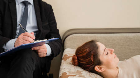 Caucasian woman lies on a couch at a reception with a psychotherapist. An elderly man works as a...