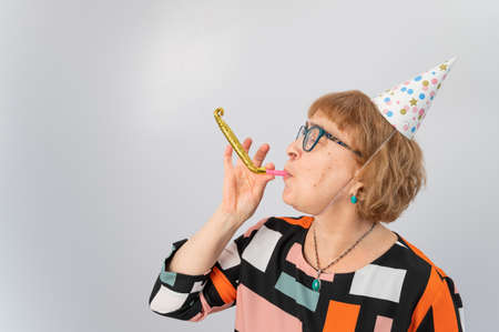 Portrait of a smiling elderly woman in a festive cap holding a whistle tongue on a white background