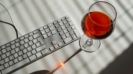 A goblet with red wine and a keyboard on a white table with shade from blindsの写真素材