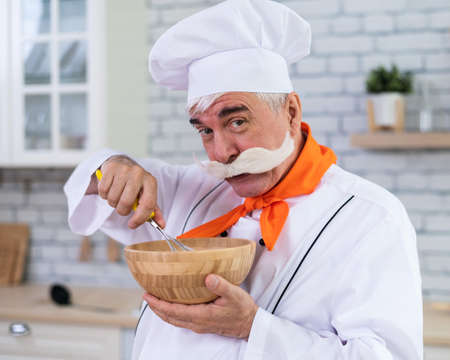 An old man with a bushy gray mustache in a chefs hat is cooking in the kitchen.の写真素材