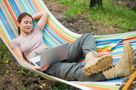 Caucasian woman working on laptop while sitting in a hammock in the forest. Girl uses a wireless computer on a hike.の写真素材