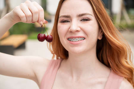Beautiful young red-haired woman with braces on her teeth eats sweet cherries in the summer outdoorsの写真素材
