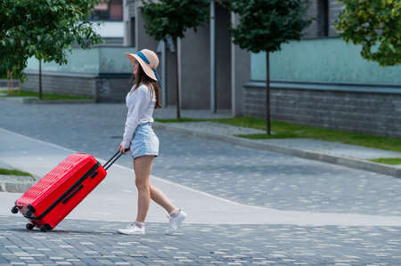 Happy caucasian young woman in a hat and shorts is holding a big red suitcase is walking on a city streetの写真素材