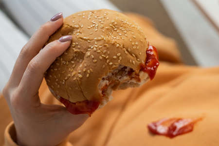 Caucasian young woman eating burger and getting dirty in ketchup. Sloppy girlの写真素材
