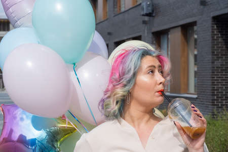 Woman in colored hair walks with an armful of balloons and drinks a refreshing beverage