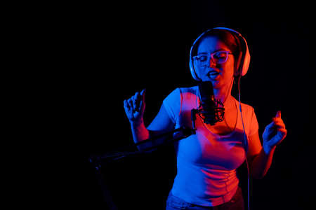 Caucasian woman in glasses and headphones sings into a microphone in neon light on a black...