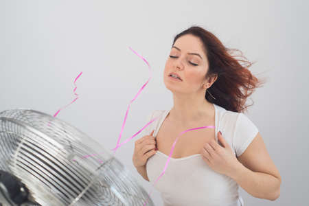 Joyful caucasian woman enjoying the wind blowing from an electric fan on a white background.