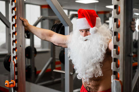 Santa claus in the gym. Muscular man with a naked torso doing exercises with dumbbells.の写真素材