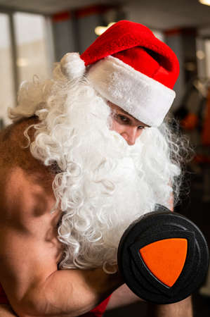Santa claus in the gym. Muscular man with a naked torso doing exercises with dumbbells.の写真素材
