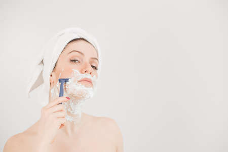 Cheerful caucasian woman with a towel on her head and shaving foam on her face holds a razor on a white backgroundの写真素材