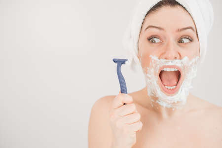 Funny portrait of a woman with shaving foam on her face holding a razor on a white background. The girl removes the mustache and beardの写真素材