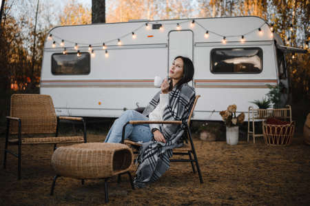 Beautiful caucasian woman is sitting wrapped in a plaid and drinking a warming drink outdoors. Traveling in a motor home in autumn.の写真素材