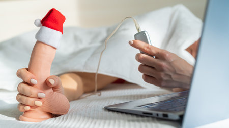 A woman lies in bed and holds a dildo with a remote control in a santa claus hat and watches porn on...