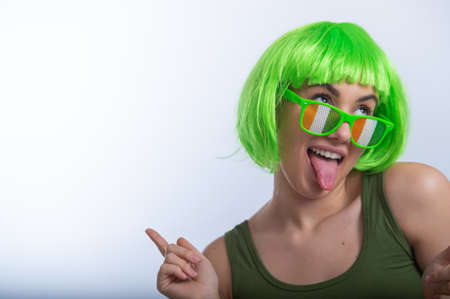 Cheerful young woman in green wig and funny glasses celebrating st patricks day on a white...