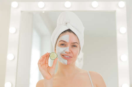 Cheerful woman with a towel on her hair and in a clay face mask fooling around with cucumbers in her hands. Taking care of beauty at homeの写真素材
