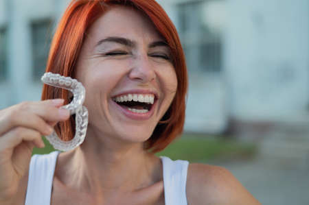 Red-haired Caucasian woman holding transparent mouthguards for bite correction outdoors. A girl with a beautiful snow-white smile uses silicone bracesの写真素材