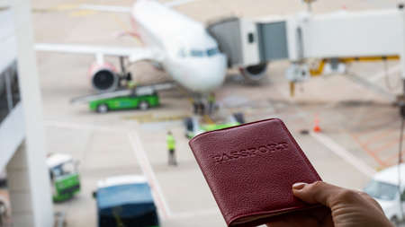 A woman holds a passport against the background of an airplane at the airport. The faceless girl is waiting for her flight and looks at the airfield through the window.の写真素材