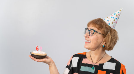A portrait of a smiling elderly woman in a festive cap holding a cake with candles in the form of...