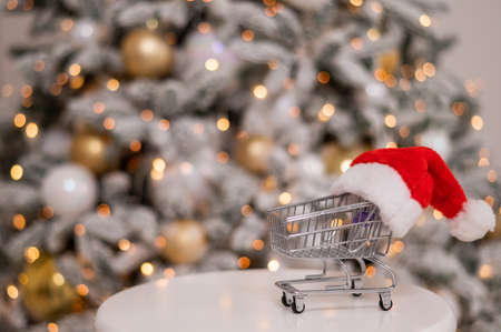 Christmas gifts shopping time. Shopping trolley in a santa claus hat by the christmas tree.