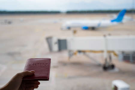 A woman holds a passport against the background of an airplane at the airport. The faceless girl is waiting for her flight and looks at the airfield through the window.の写真素材