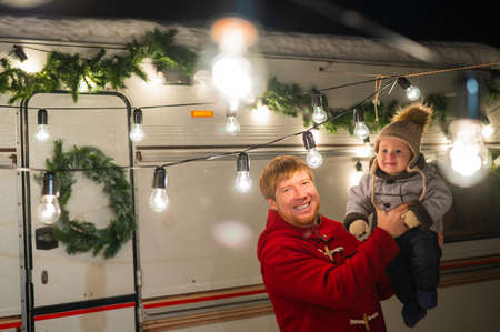 Caucasian red-haired man with a boy in his arms at the mobile home. Father and son celebrate Christmas on a trip.の写真素材