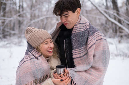 A young couple walks in the park in winter. The guy and the girl are drinking a warming drink...