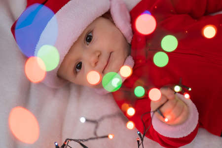 A baby boy dressed in a santa suit lies on the bed and plays with a Christmas garland.