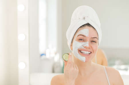 Cheerful woman with a towel on her hair and in a clay face mask fooling around with cucumbers in her...
