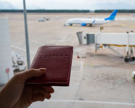 A woman holds a passport against the background of an airplane at the airport. The faceless girl is waiting for her flight and looks at the airfield through the window.の写真素材