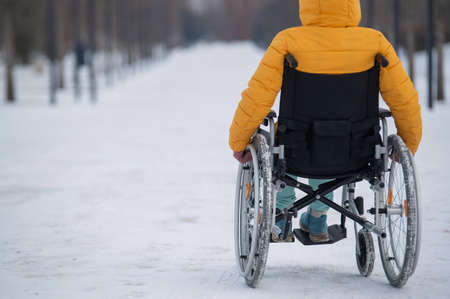 Caucasian woman with disabilities rides on a chair in the park in winter. Back view of a girl on a walk in a wheelchair.の写真素材