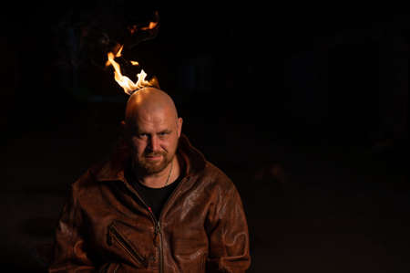 A bald man in a leather jacket with a burning head grins.