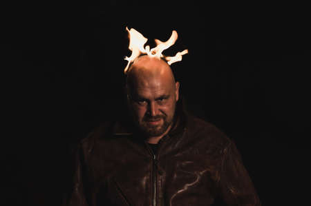 A bald man in a leather jacket with a burning head grins.