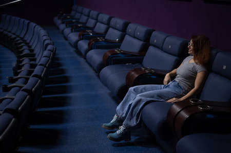 Caucasian red-haired woman sits on the last row in a cinema in an empty hall. The girl is watching a...