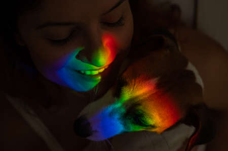 Close-up portrait of a woman hugging a jack russell terrier dog with a ray of rainbow light on her face.の写真素材