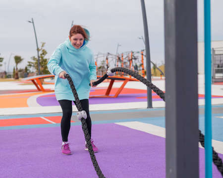 Caucasian woman in a mint sweatshirt is training with battle ropes at the sports ground.の写真素材
