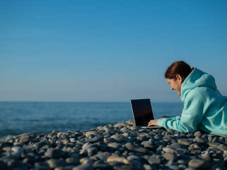 Caucasian woman lying on her stomach on a pebble beach and typing on a laptop. Remote work freelancer.の写真素材