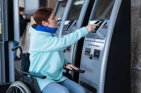Caucasian woman in a wheelchair buys a train ticket using a mobile phone at a self-service checkout.の写真素材