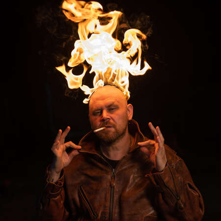 Bald man in a leather jacket with a burning head on a dark background.