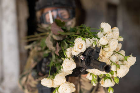 Caucasian woman in military uniform holding a machine gun and a bouquet of white roses.